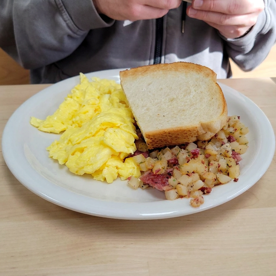 Plate of scrambled eggs, toast, and corned beef hash on a wooden restaurant table, with a diner seated behind it.